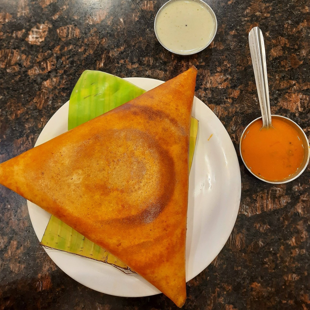 A crisp golden dosa folded into a triangle on a banana leaf, with coconut chutney and sambar