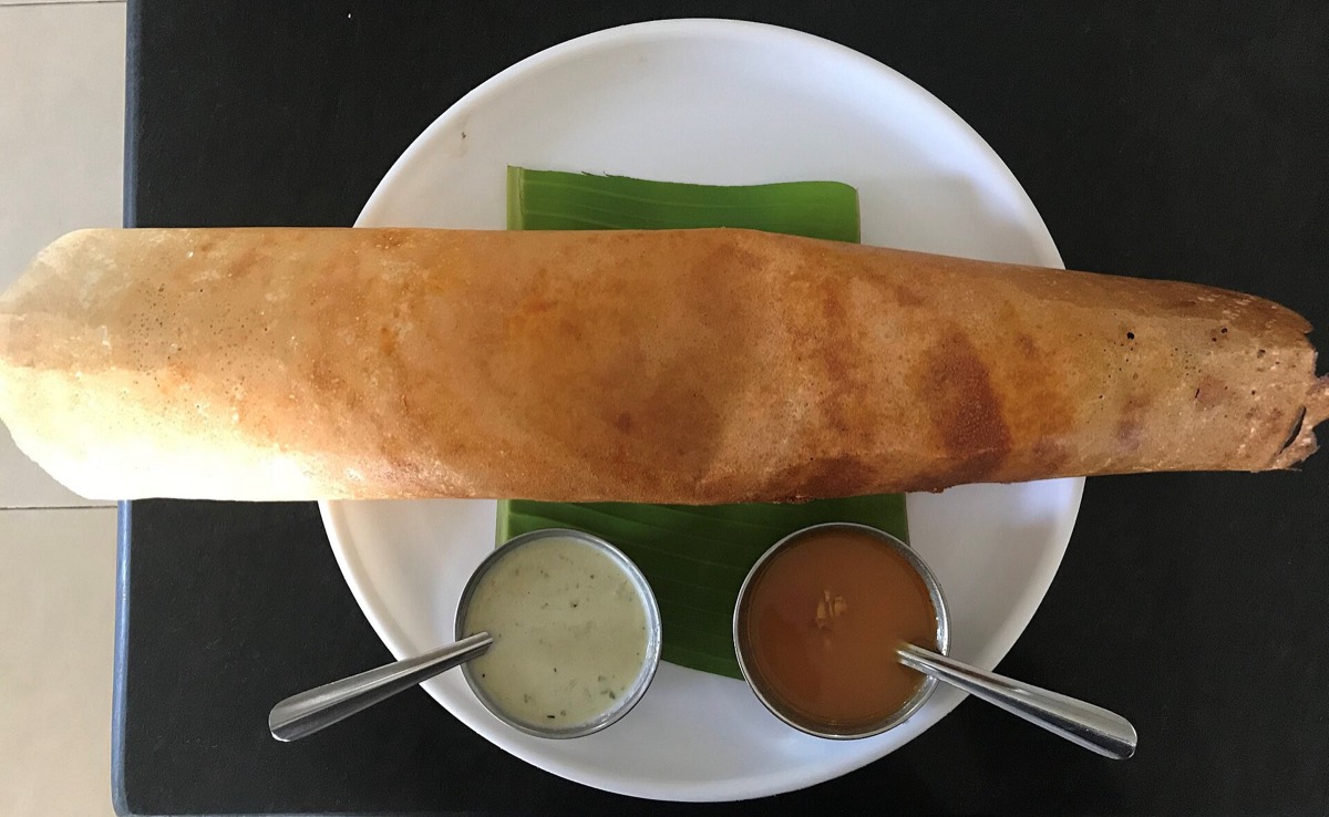 A long rolled masala dosa on a banana leaf with chutney and sambar